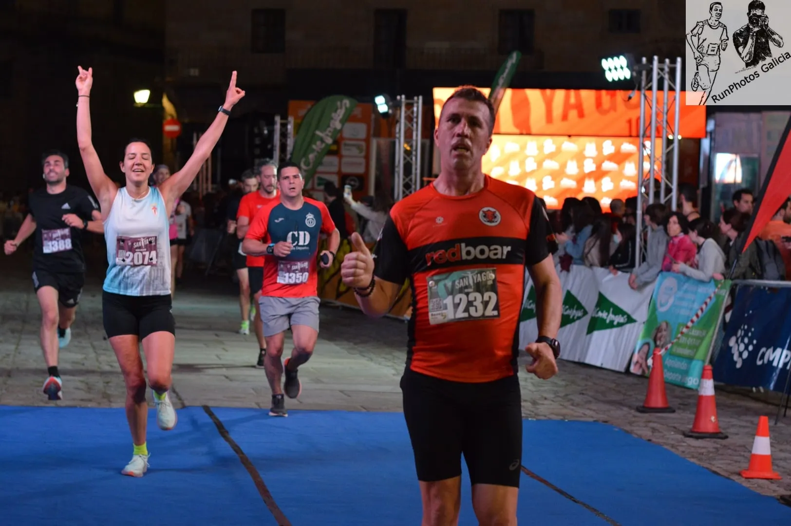 Runner giving thumbs up crossing finish line with celebratory runner behind and crowd of spectators watching