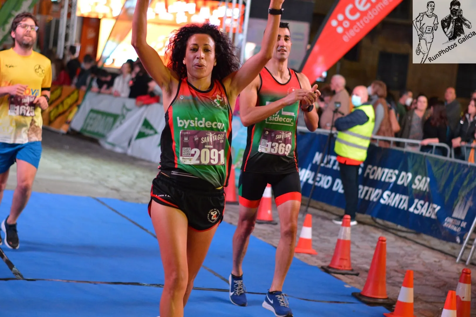 Female runner celebrating with arms raised as she crosses the finish line on blue mat with spectators cheering
