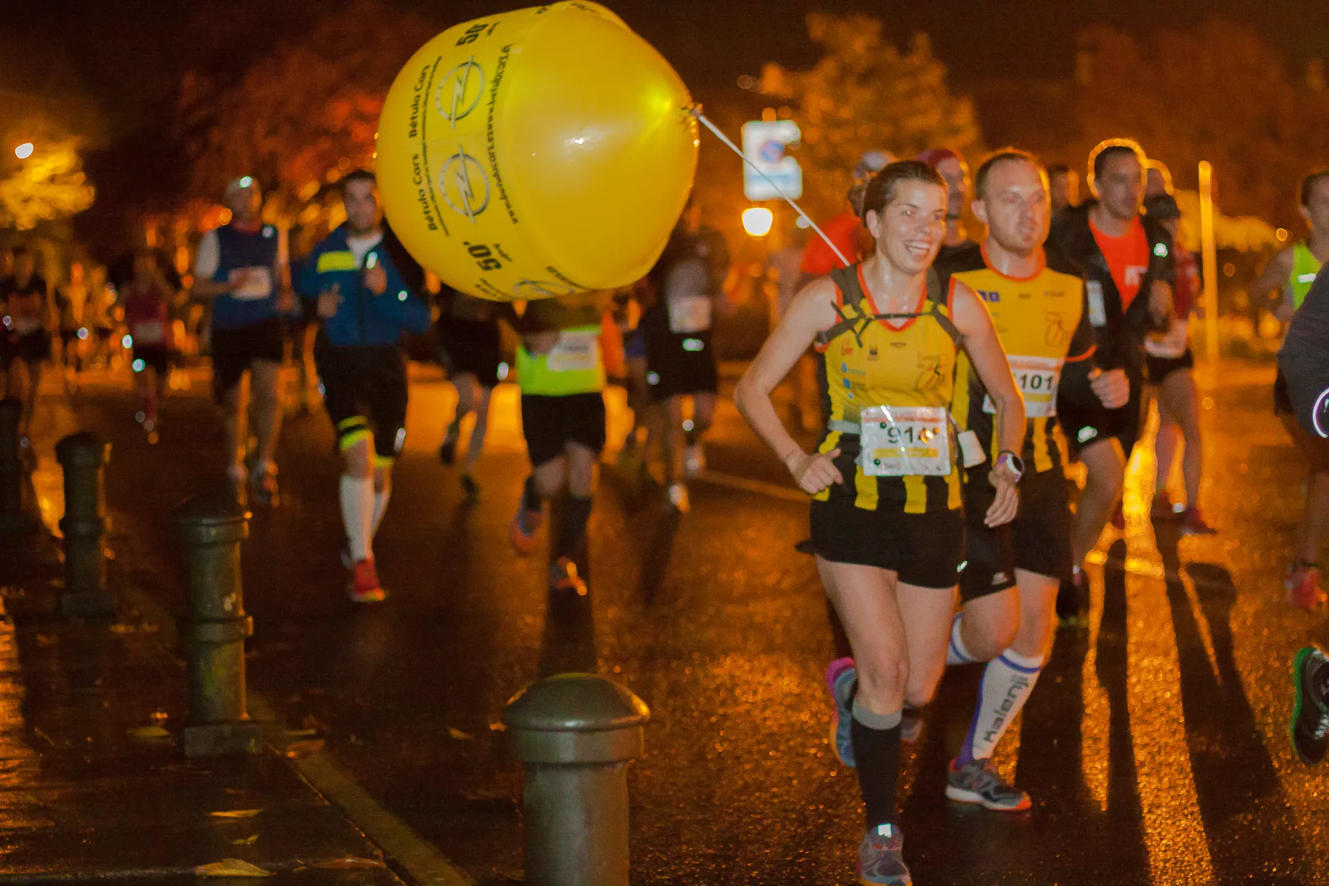 Smiling runner holding large yellow balloon while running through wet night streets with other participants