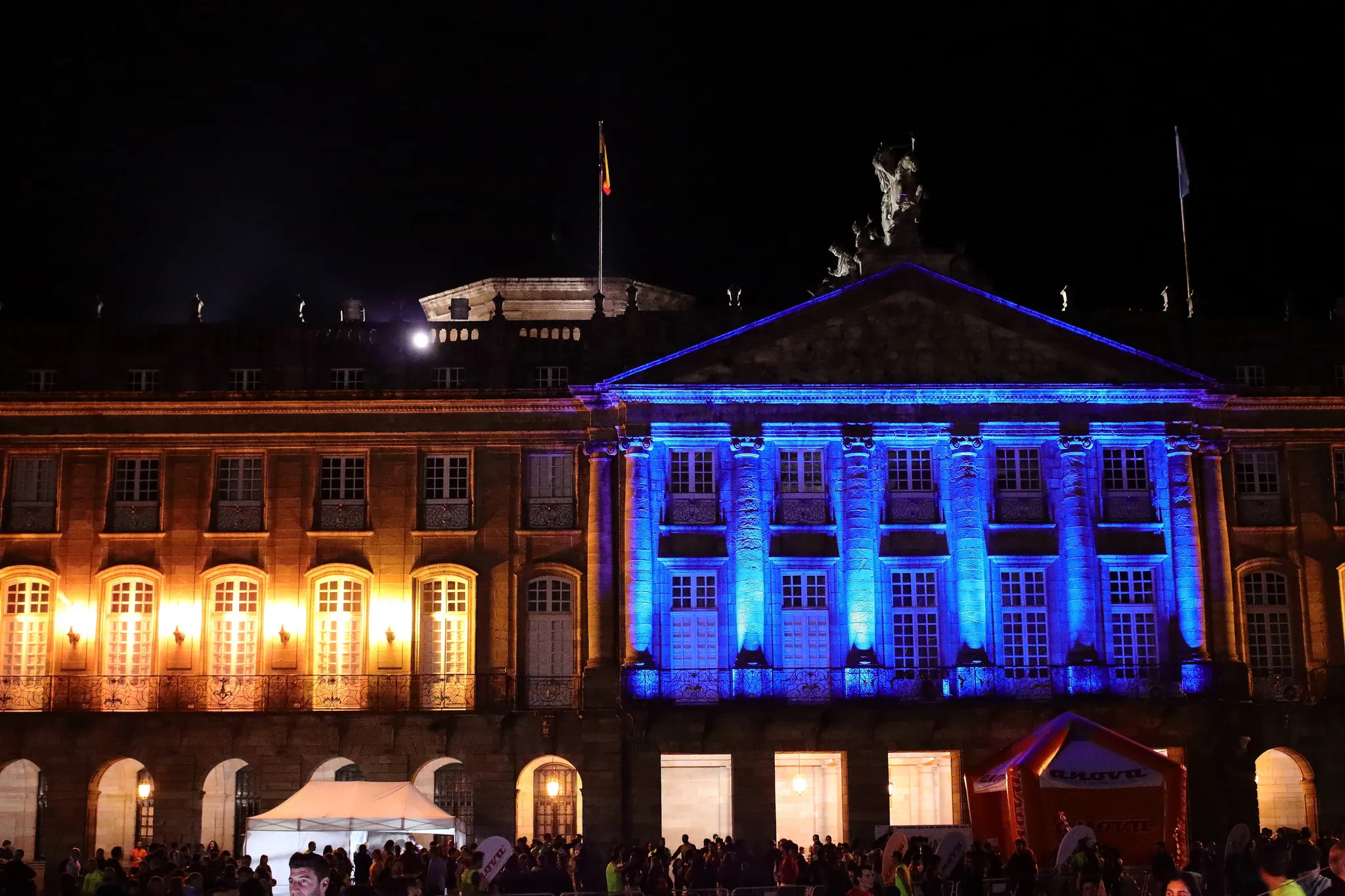 Rajoy Palace in Obradoiro Square illuminated with blue and orange lights at night with race venue below