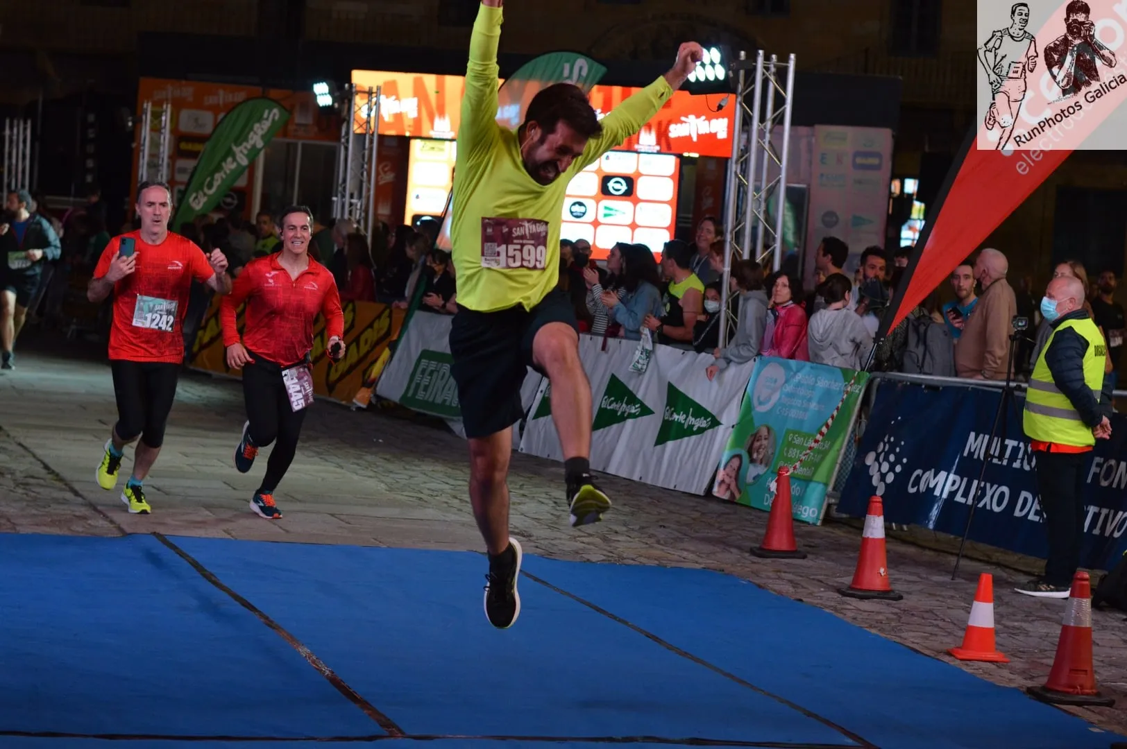 Runner in bright yellow shirt jumping for joy with arms raised as he crosses the finish line with crowd behind