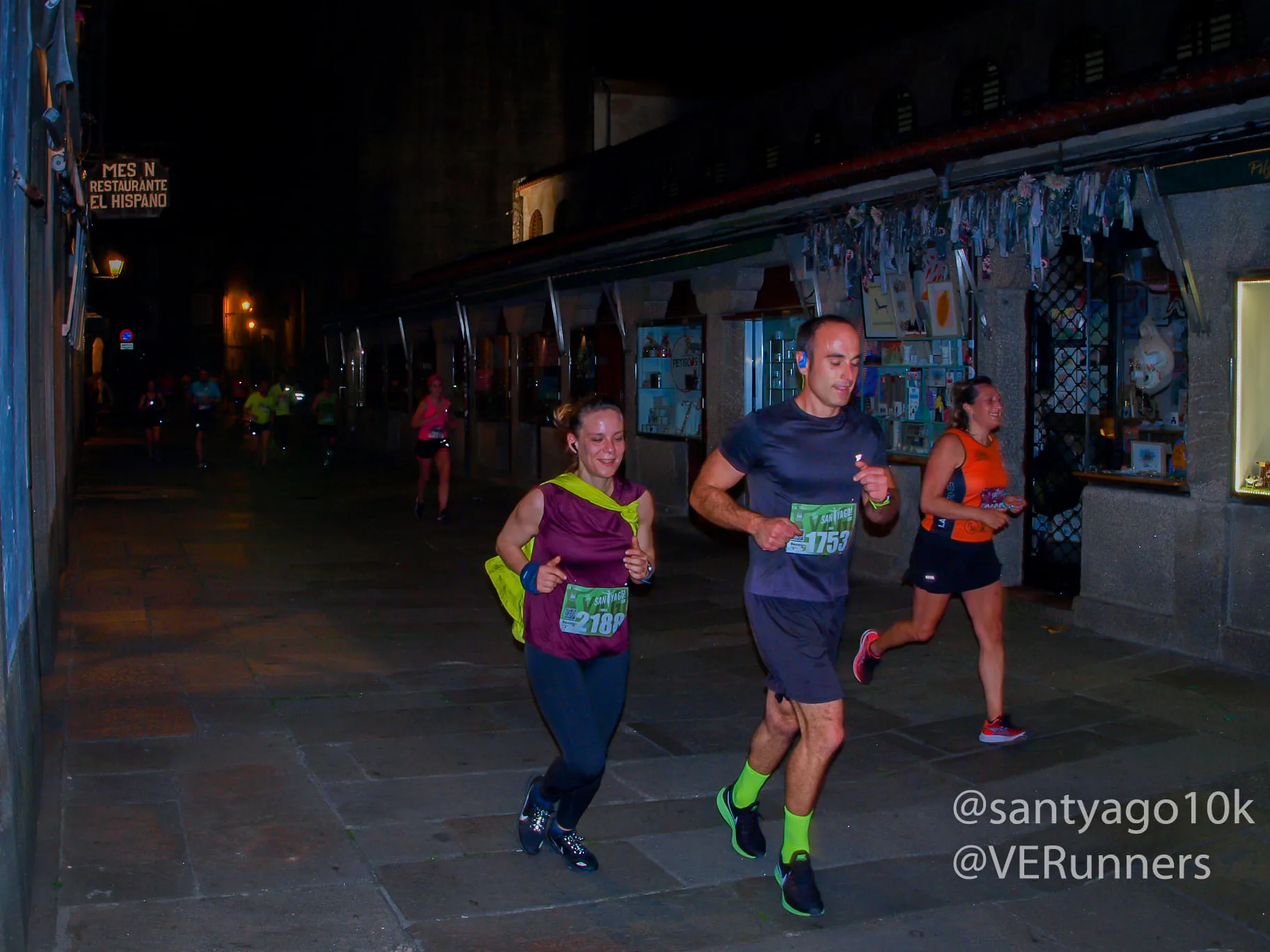 Small group of runners passing through narrow cobblestone streets past historic storefronts at night
